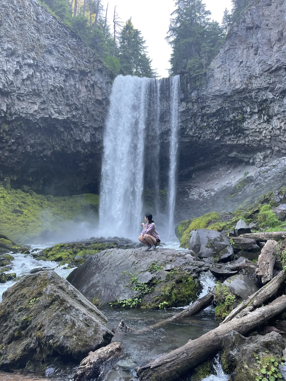 Michelle crouched on a mossy boulder at the base of a tall waterfall, looking up at the falling water in a basalt canyon lined with moss and fallen logs